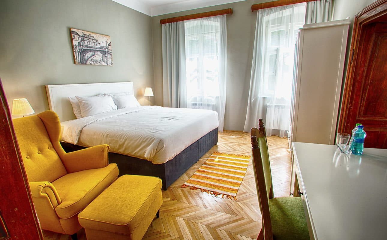 The standard double room, with a double bed, a yellow armchair and footstool, and a striped runner on herringbone parquet.