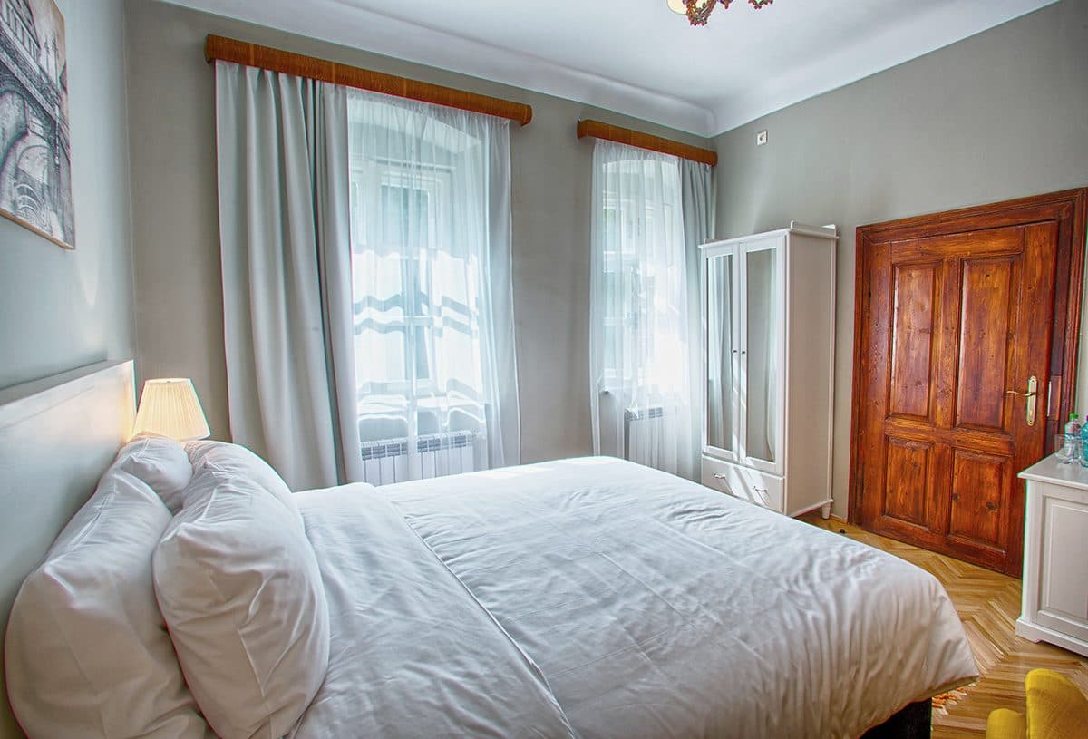 The bed in the standard double room, beside the curtained window and a bedside lamp.