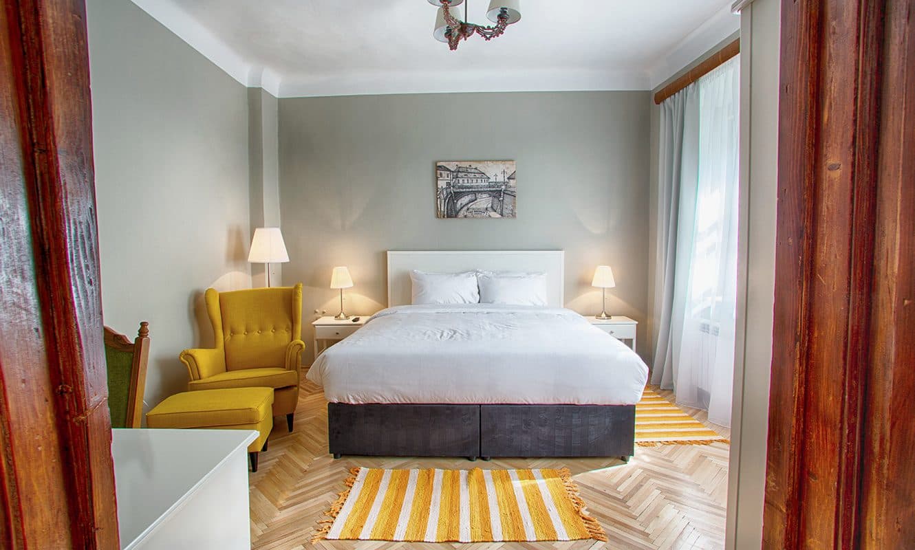 The standard double room seen from the doorway, with the yellow armchair, the bed and the tall wooden door.