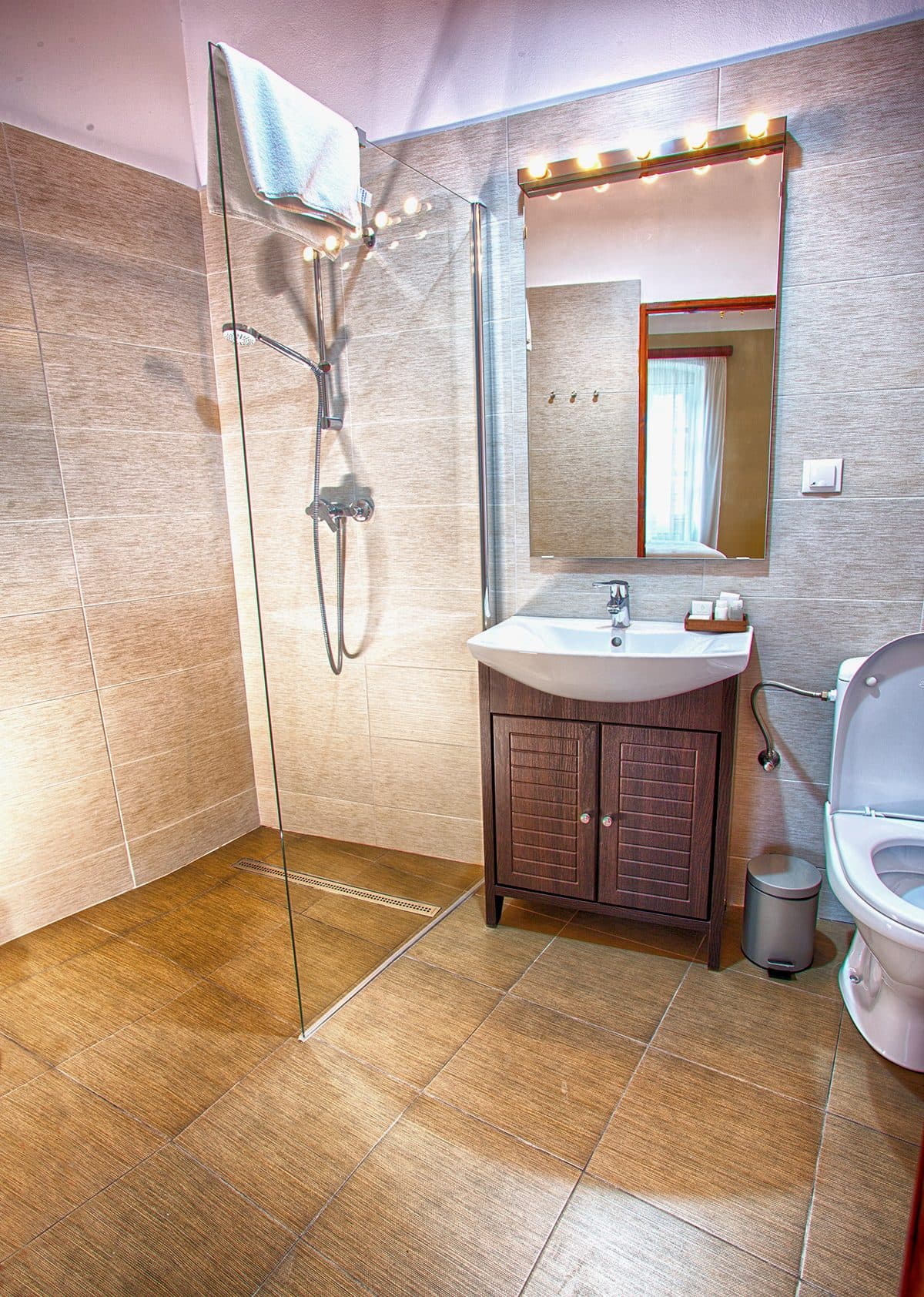 A superior room's bathroom, with a shower, a basin set on wooden furniture and a mirror with bulbs.