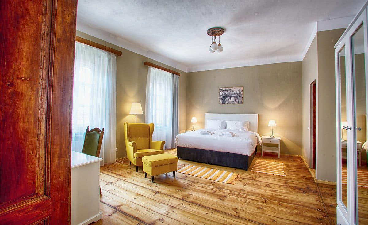 A superior room, with a tall wooden door, a double bed and a yellow armchair by the window.