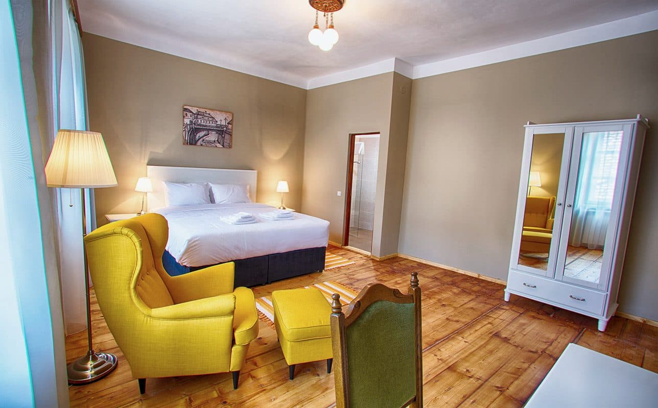 A superior room, with a yellow armchair, footstool, the bed and the door to the hall.