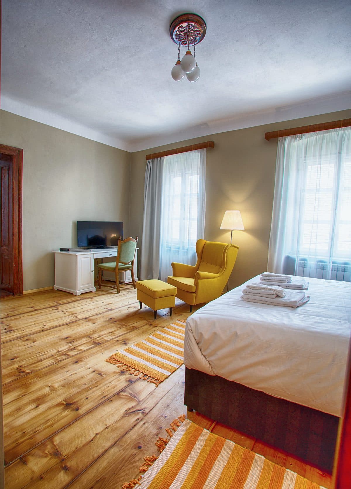 A superior room, with a television, armchair and bed, under a high ceiling with a light.