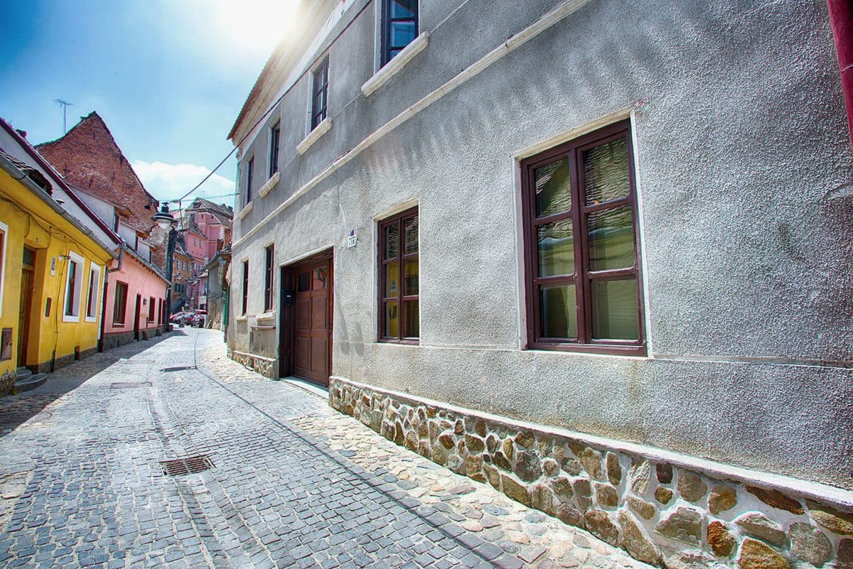 The house's grey rendered facade on Strada Târgului, with its river-stone plinth, red-brown timber windows and the large wooden gate, on a sloping cobbled street.
