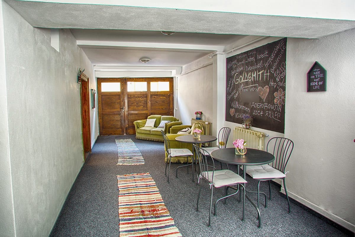 The vaulted passage between the gate and the courtyard, with woven runners on the floor, olive-green sofas, bistro tables, and the large writing board chalked with GOLDSMITH surrounded by welcomes in many languages.
